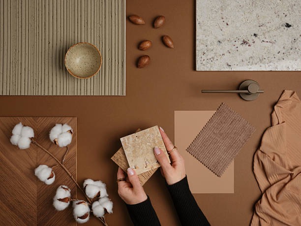 Paleta barw i materiałów do aranżacji wnętrz w odcieniach brązu, z dłońmi trzymającymi próbki płytek. Widoczne drewno, ceramika, tkanina i element armatury.