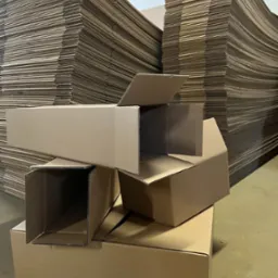 Stos kartonowych pudeł i płaskich arkuszy tektury falistej w magazynie. Pudełka różnej wielkości, niektóre otwarte, ukazują strukturę kartonu. Umiarkowane oświetlenie, surowy klimat.