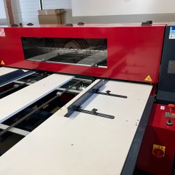 BOX-MASTER - Czerwono-szara maszyna do pakowania z widocznymi elementami transportu wewnętrznego i panelami sterowania. Urządzenie przemysłowe w hali produkcyjnej.