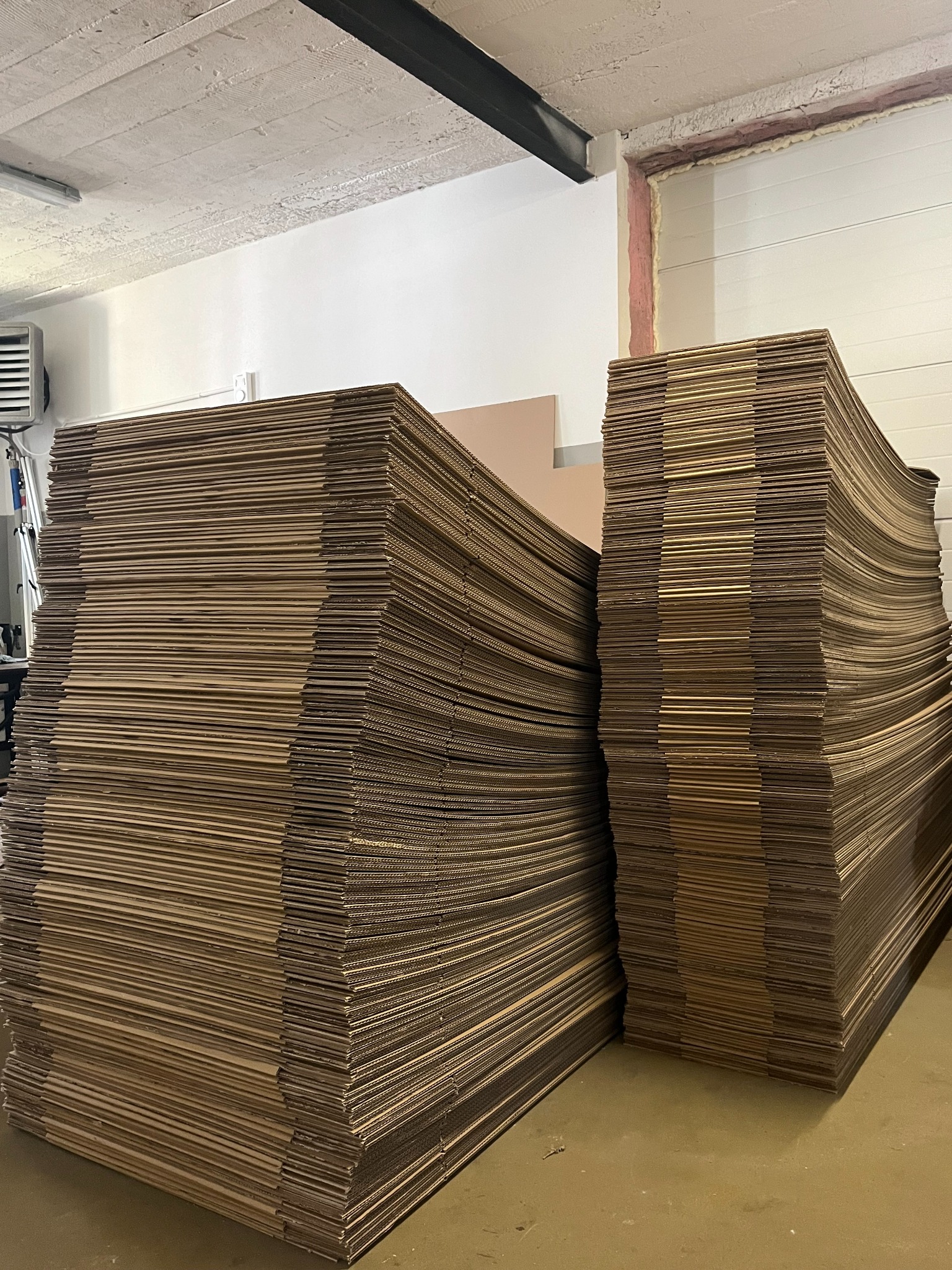 Dwa stosy płaskich, brązowych arkuszy tektury falistej w magazynie, ustawione jeden za drugim, z widoczną strukturą i grubością warstw. Oświetlenie magazynowe.
