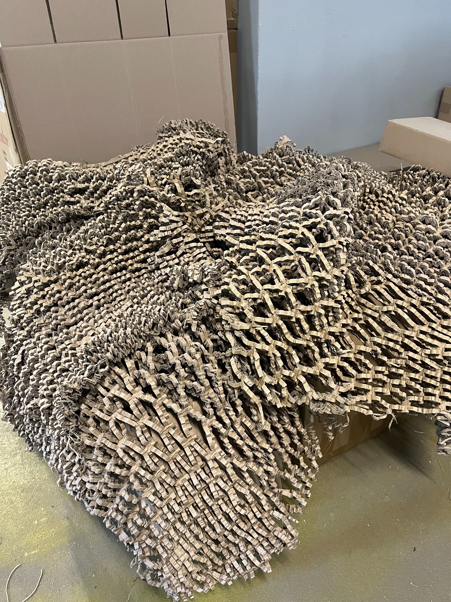 Stos brązowego, perforowanego papieru naciętego w kratkę, używanego jako wypełniacz do paczek, na tle kartonowych pudeł i szarej ściany.