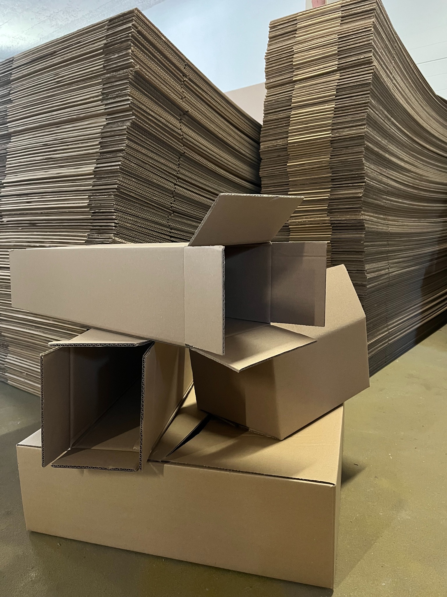 Stos kartonowych pudeł i płaskich arkuszy tektury falistej w magazynie. Pudełka różnej wielkości, niektóre otwarte, ukazują strukturę kartonu. Umiarkowane oświetlenie, surowy klimat.