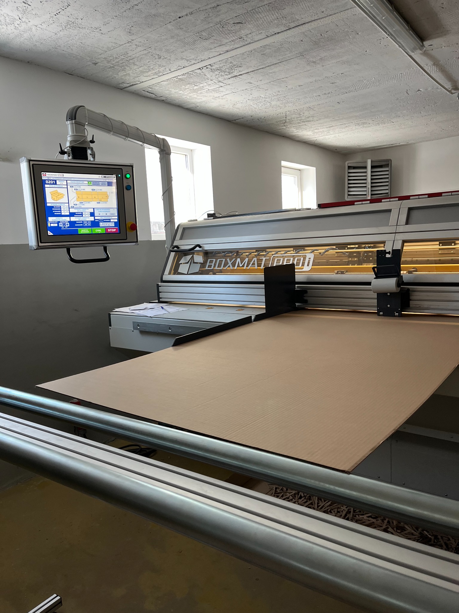 Automatyczna maszyna BOXMAT PRO do cięcia kartonu, z ekranem dotykowym i arkuszem tektury na stole roboczym. Widoczne oświetlenie i elementy konstrukcyjne maszyny.