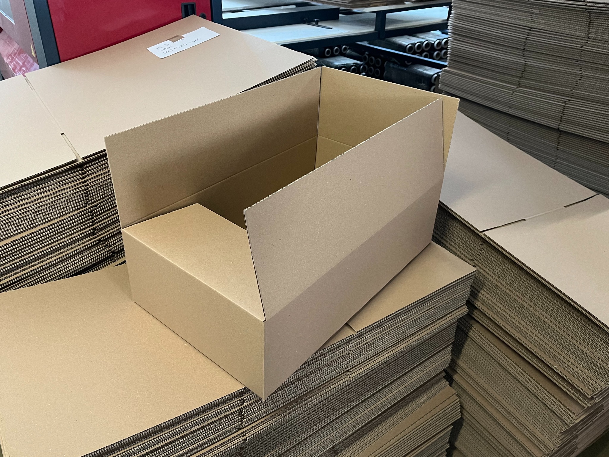 Rozłożony kartonowy pojemnik na stosie płaskich arkuszy, gotowy do złożenia. Widoczne wymiary na jednym z arkuszy. Tło stanowią elementy linii produkcyjnej.