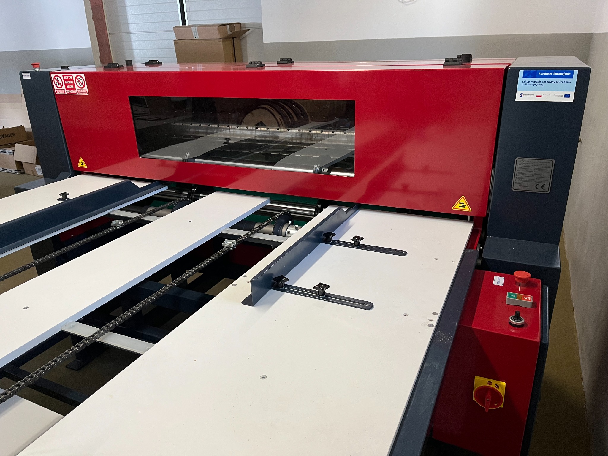 Czerwono-szara maszyna do pakowania z widocznymi elementami transportu wewnętrznego i panelami sterowania. Urządzenie przemysłowe w hali produkcyjnej.