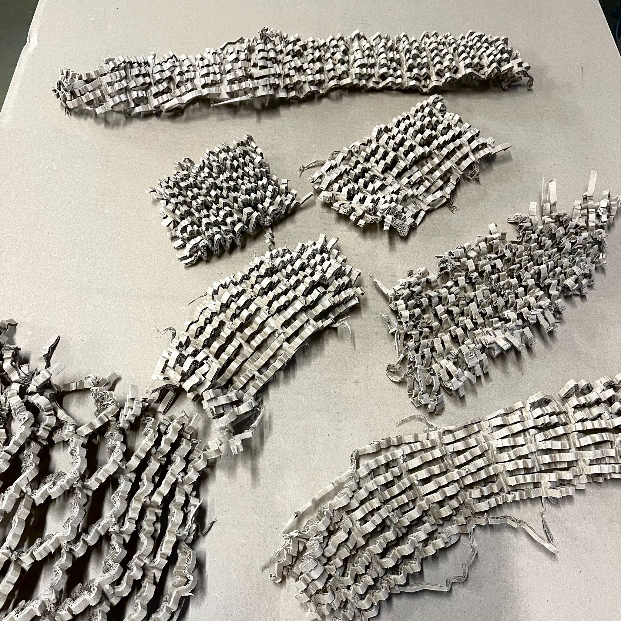 Falista tektura pocięta na paski, ułożona w nieregularne kształty na szarym tle. Materiał wypełniający paczki, widok z góry, oświetlenie studyjne.