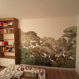 AAPARBUILD - Pokój po malowaniu z fototapetą leśną na ścianie. Widoczny regał z książkami i fragment łóżka. Gniazdka elektryczne wkomponowane w motyw drzew.