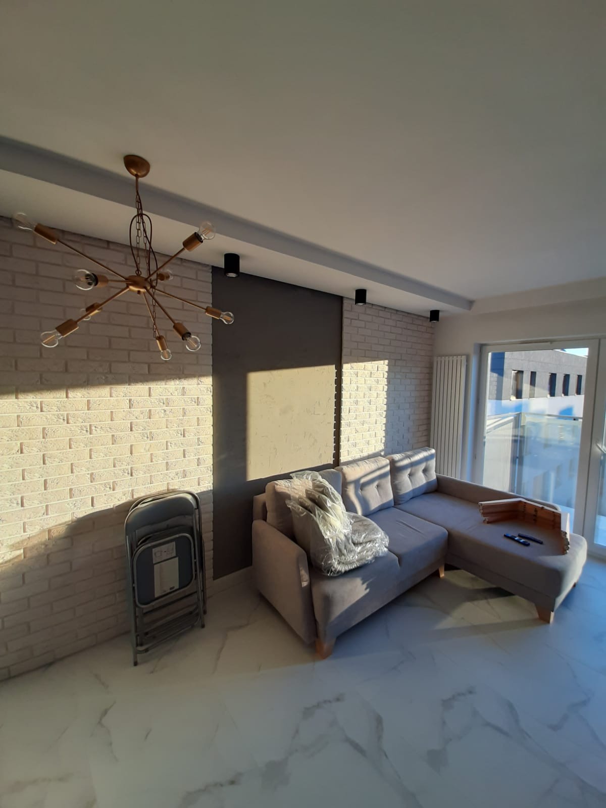 Jasne wnętrze po remoncie: sofa z poduszkami w folii, ściana z cegły, designerska lampa i marmurowe płytki podłogowe. Widoczne efekty malowania i wykończenia.