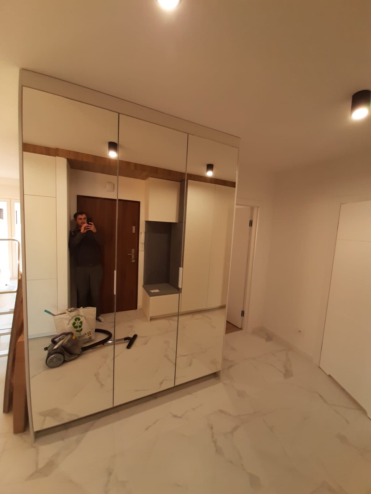 Nowoczesna zabudowa przedpokoju z lustrzanymi drzwiami przesuwnymi, odbijająca wnętrze mieszkania i osobę robiącą zdjęcie. Białe fronty szaf i marmurowe płytki podłogowe.