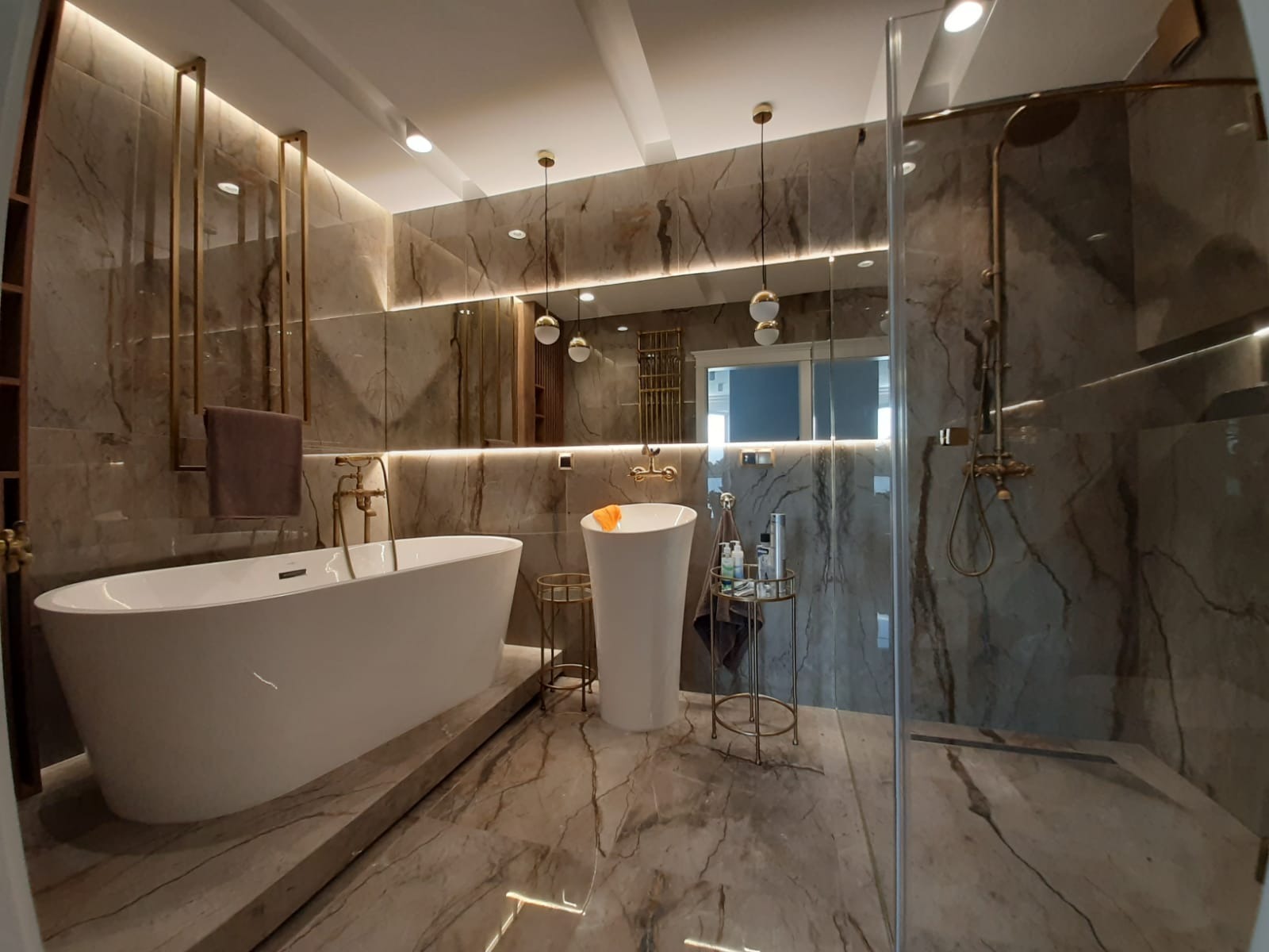 Elegancka łazienka w odcieniach beżu i złota. Wolnostojąca wanna, umywalka na postumencie, kabina prysznicowa ze złotą armaturą. Marmurowe płytki i oświetlenie LED.