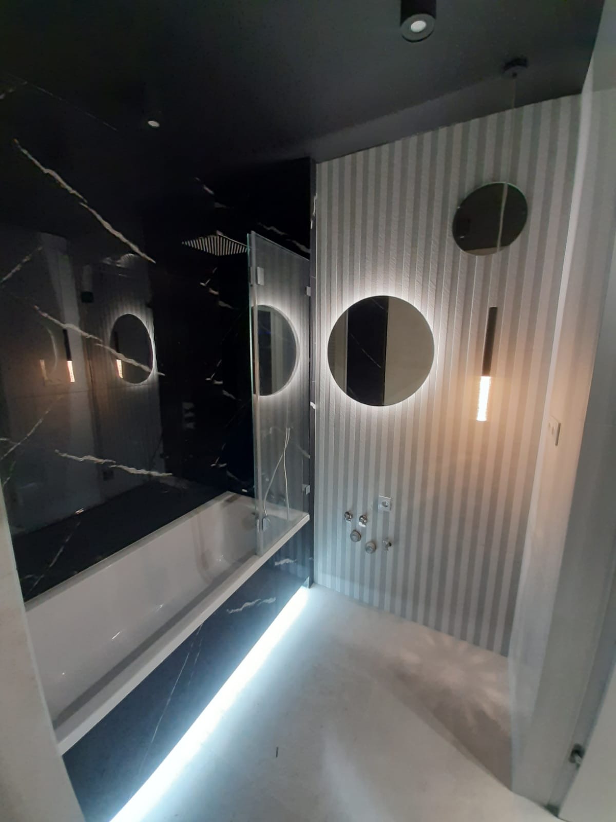 Nowoczesna łazienka z czarnymi marmurowymi płytkami, podświetlaną wanną i okrągłymi lustrami na ścianie w paski. Minimalistyczny design z akcentami światła.