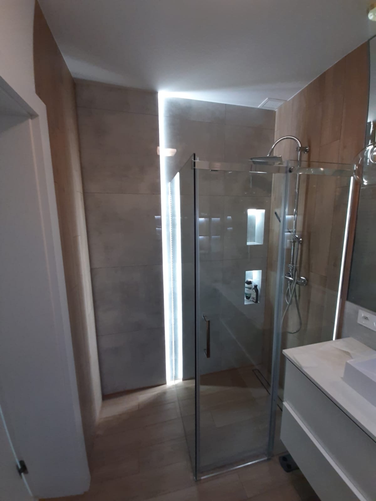 Nowoczesna kabina prysznicowa ze szklanymi drzwiami i oświetleniem LED w wykończonej łazience z drewnopodobnymi płytkami i wnękami na kosmetyki.