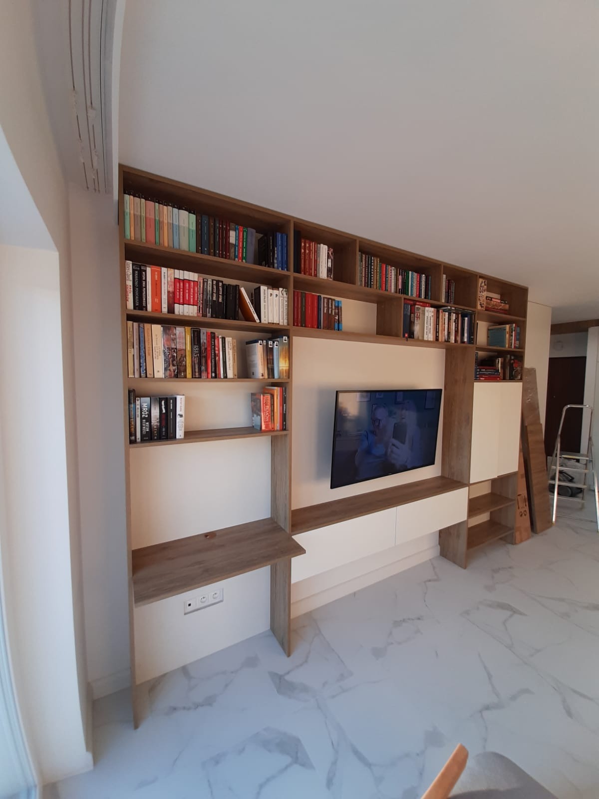 Zabudowa ściany z półkami na książki i szafkami RTV w salonie. Jasne drewno i białe fronty, marmurowa podłoga. Widoczny fragment drabiny.