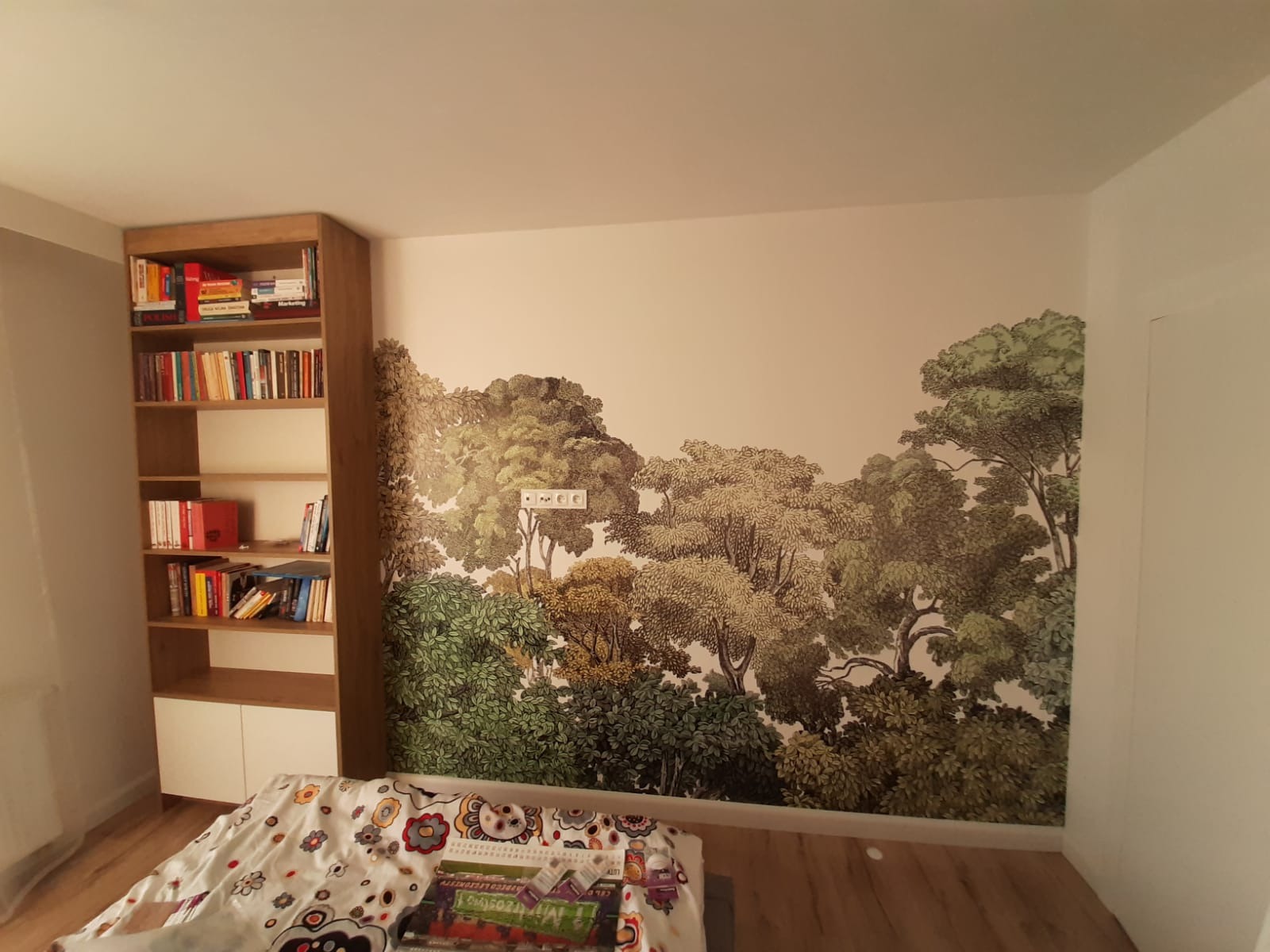Pokój po malowaniu z fototapetą leśną na ścianie. Widoczny regał z książkami i fragment łóżka. Gniazdka elektryczne wkomponowane w motyw drzew.