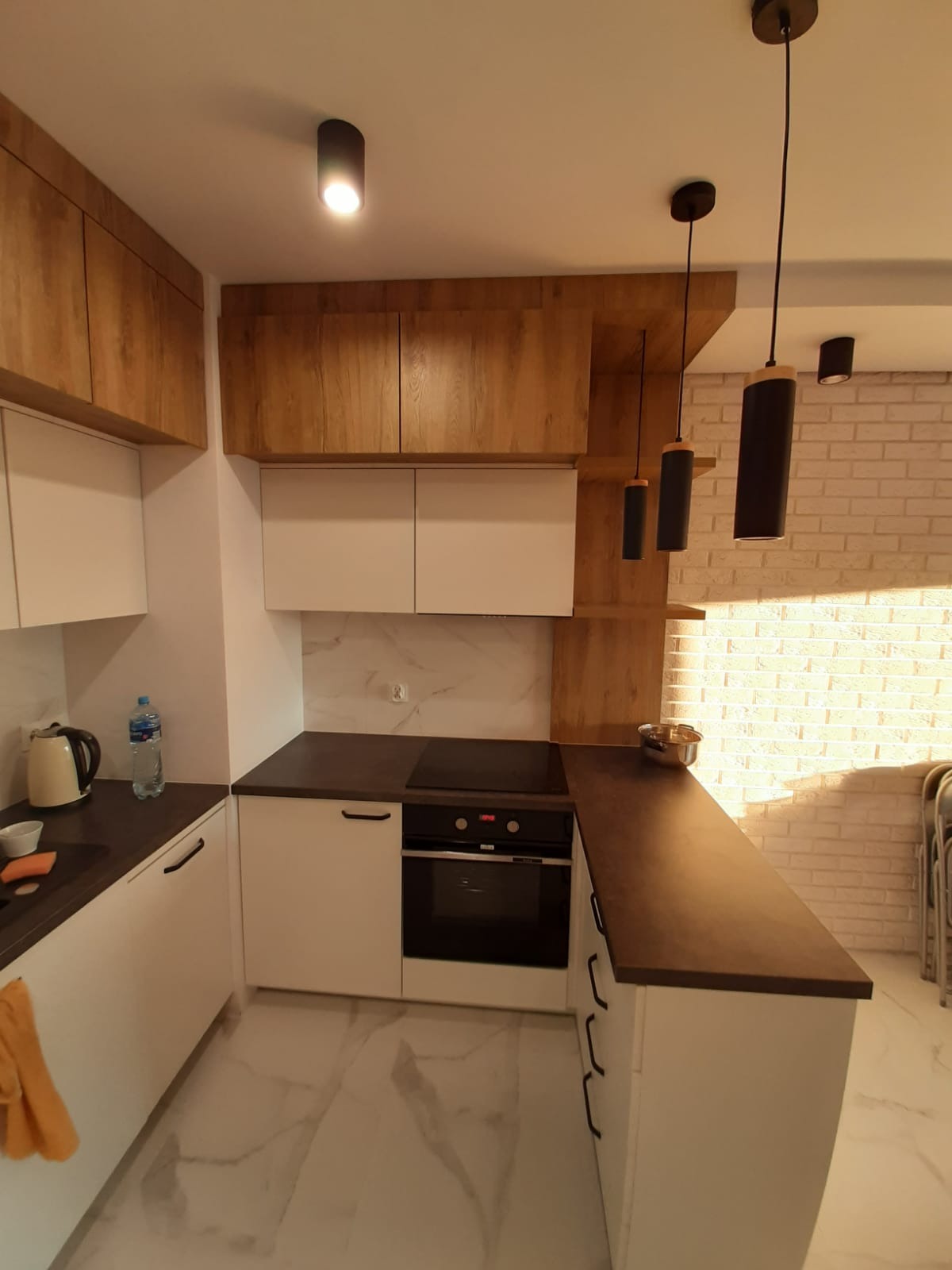 Nowoczesna kuchnia z białymi szafkami i drewnianymi akcentami, oświetlona wiszącymi lampami. Biała cegła na ścianie dodaje wnętrzu charakteru.