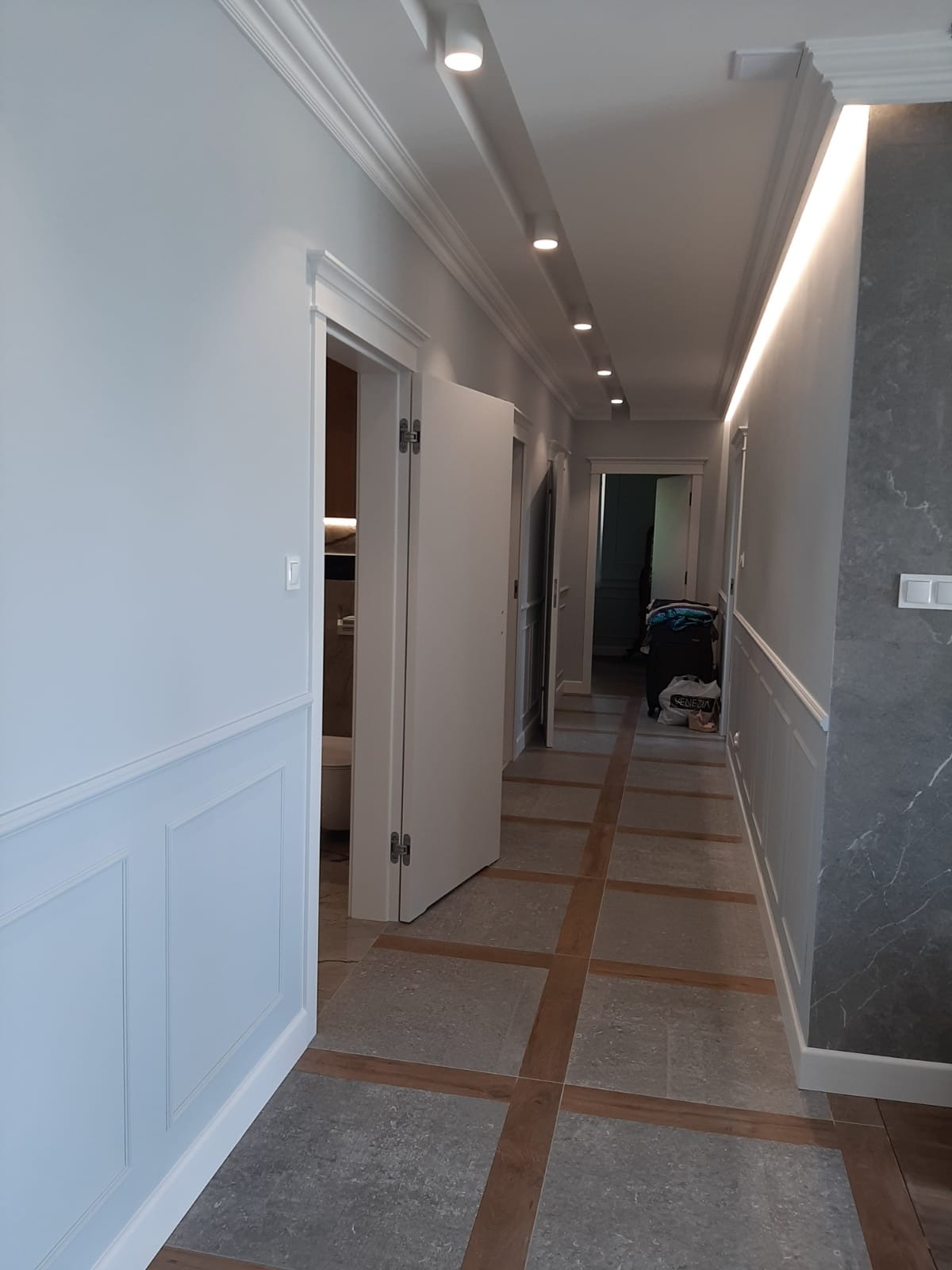 Wykończony korytarz w jasnych barwach z panelami ściennymi, oświetleniem punktowym i podłogą w kratkę. Widoczne otwarte drzwi i fragment łazienki.