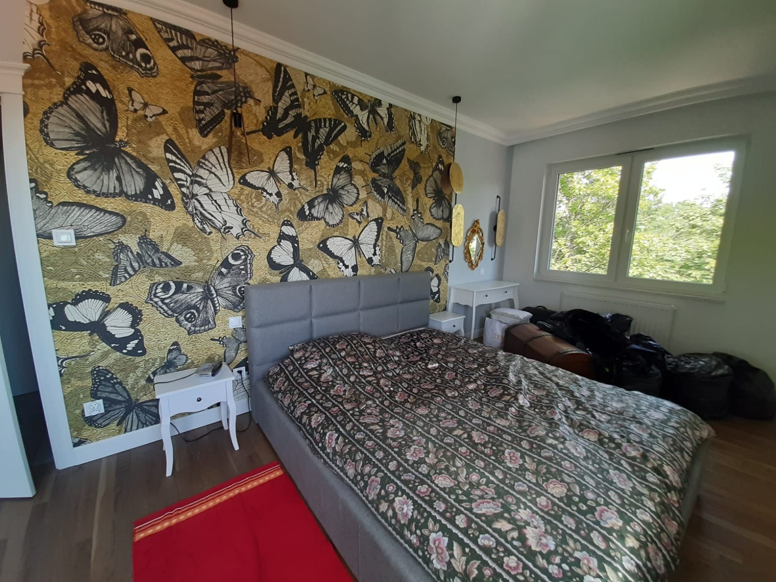 Sypialnia z łóżkiem i ścianą w złote motyle. Widoczne stoliki nocne i okno z widokiem na zieleń. Dekoracyjne lustra na ścianie. Uwagę przykuwa wzorzysta tapeta.