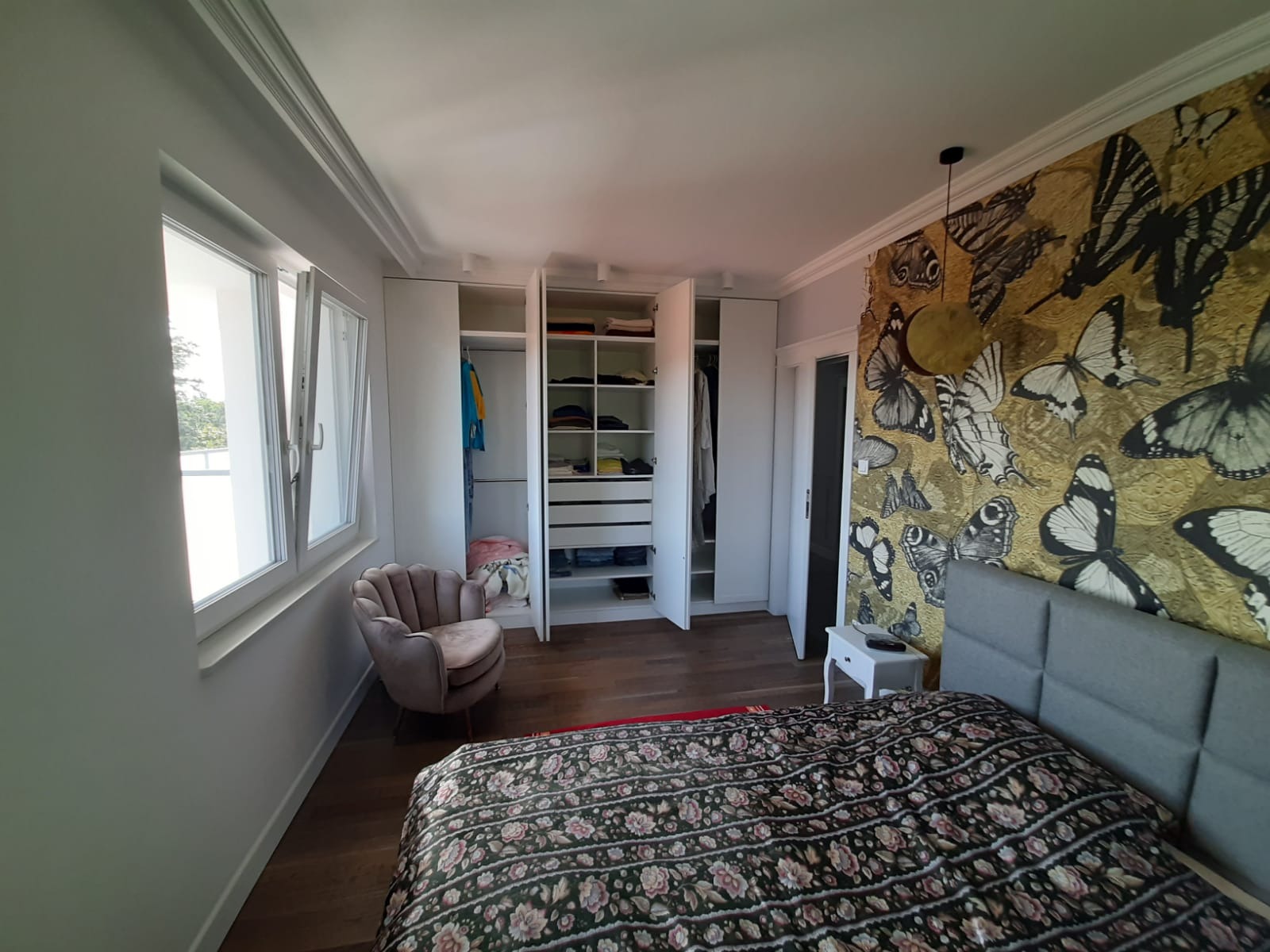 Nowoczesna sypialnia z dużą szafą wnękową z otwartymi drzwiami, parkietem i tapetą w motyle. Widok na okno i fotel. Jasne wnętrze.