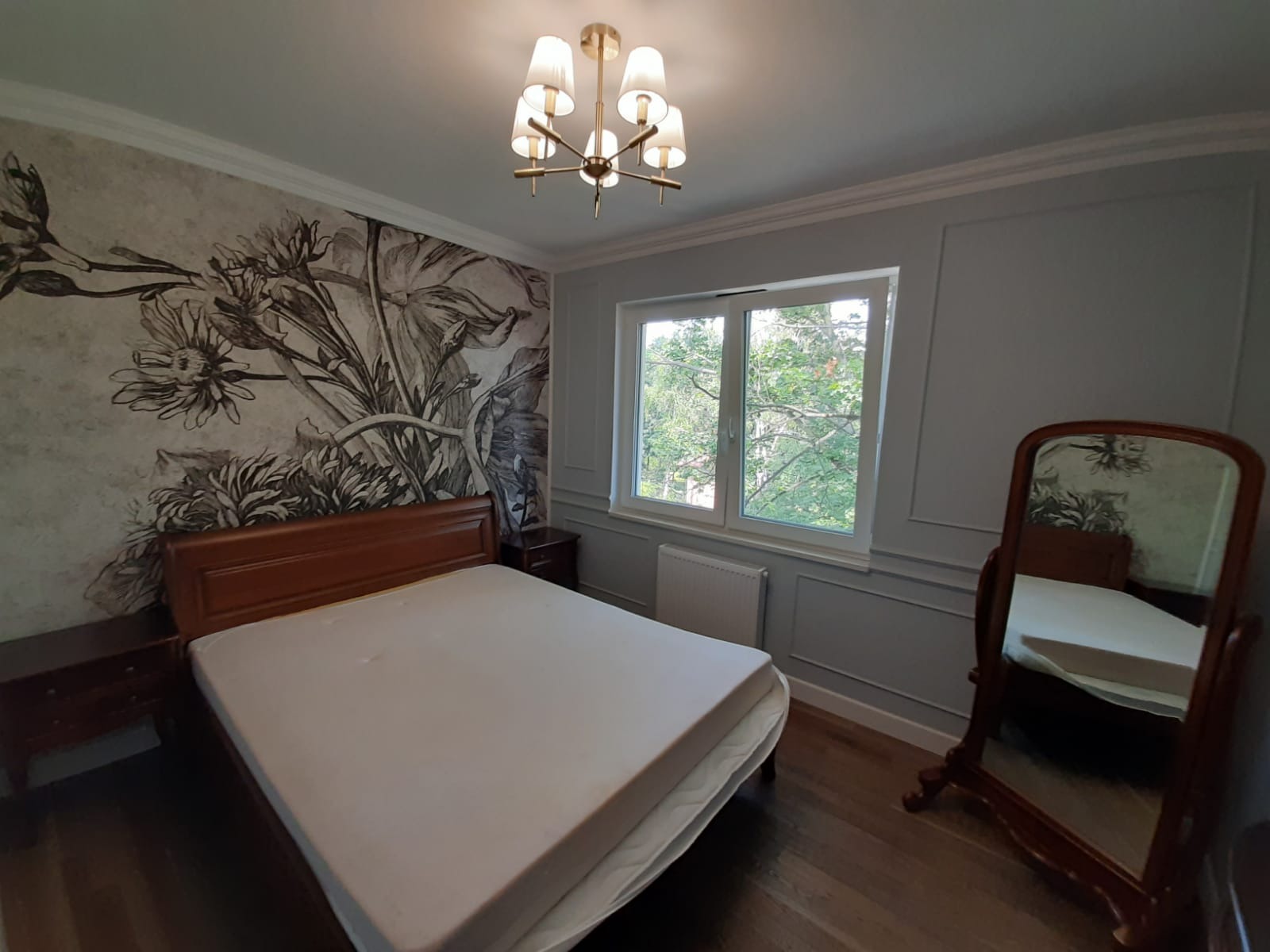 Sypialnia po remoncie: łóżko z drewnianą ramą, ściana z motywem kwiatowym, okno z widokiem na zieleń, lustro w drewnianej ramie i złoty żyrandol.