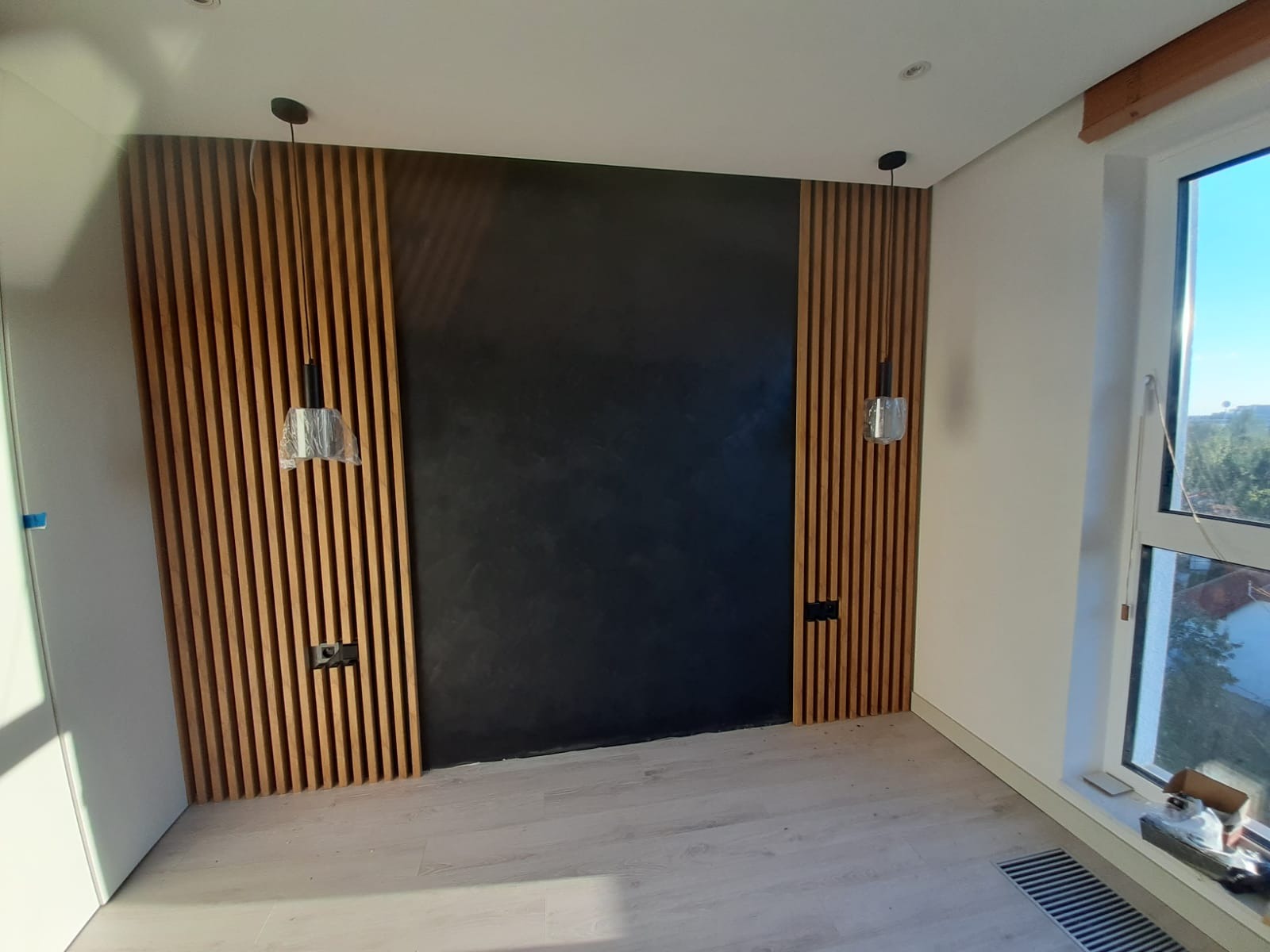 Nowoczesna ściana akcentująca z drewnianymi lamelami po bokach i czarną, teksturowaną powierzchnią pośrodku. Wiszące lampy i gniazdka elektryczne wkomponowane w konstrukcję.