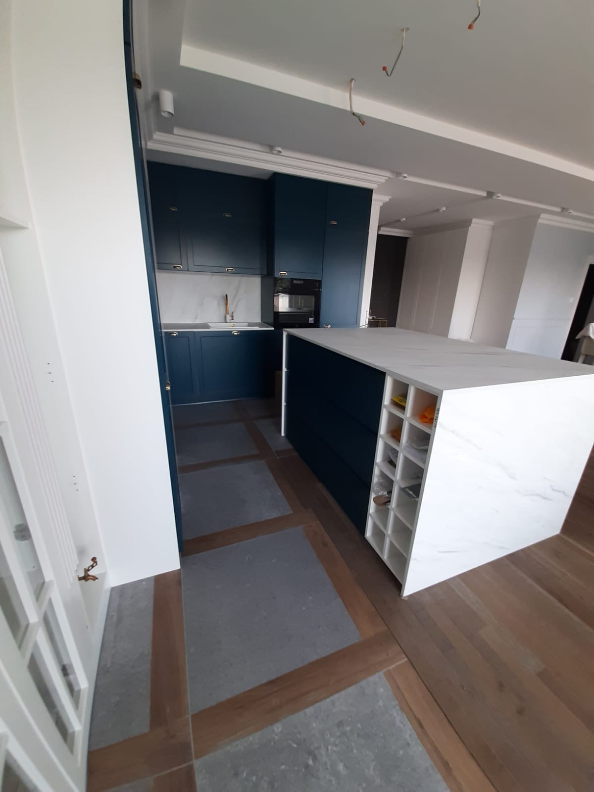 Granatowa zabudowa kuchenna z wyspą na tle marmurowej ściany. Podłoga z szarych płyt i drewnianych wstawek. Widok zza białych drzwi.