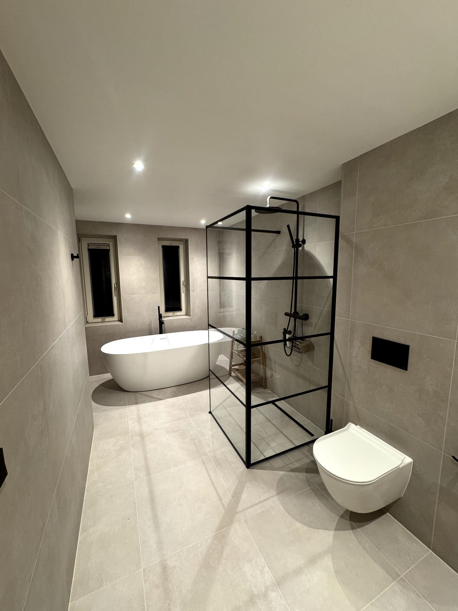 Nowoczesna łazienka w odcieniach szarości z wolnostojącą wanną, kabiną prysznicową typu walk-in z czarnymi ramami i podwieszaną toaletą. Minimalistyczny design.