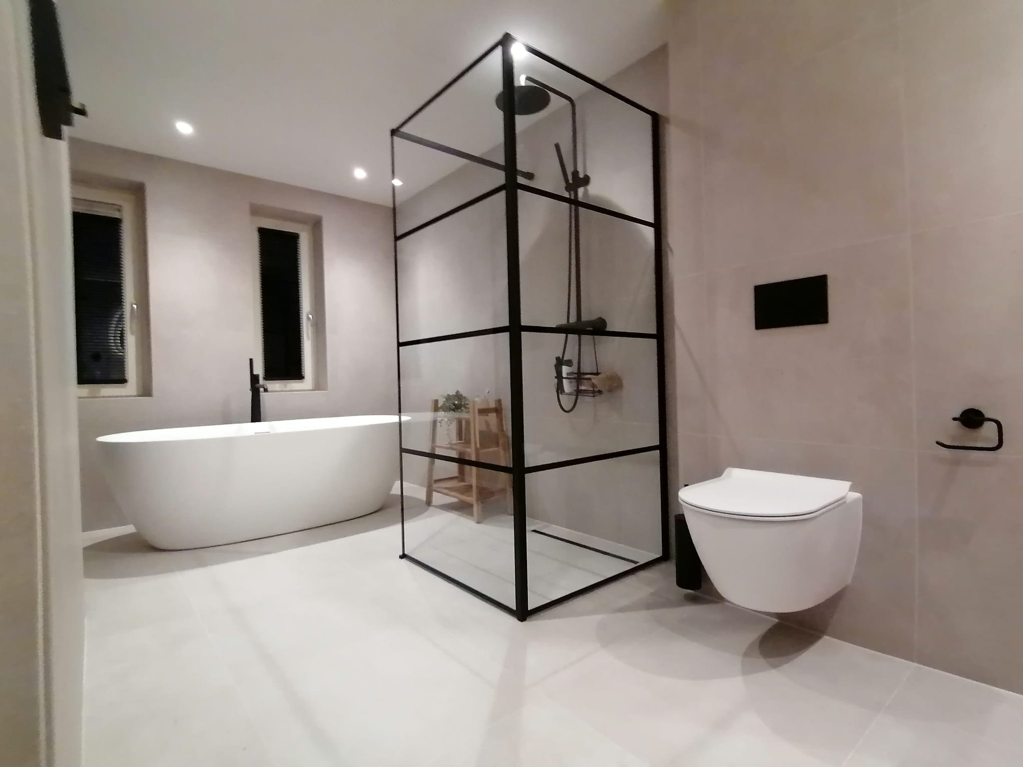 Nowoczesna łazienka z wolnostojącą wanną, kabiną prysznicową typu walk-in z czarnymi profilami i podwieszaną toaletą. Minimalistyczny design, jasne płytki, punktowe oświetlenie.