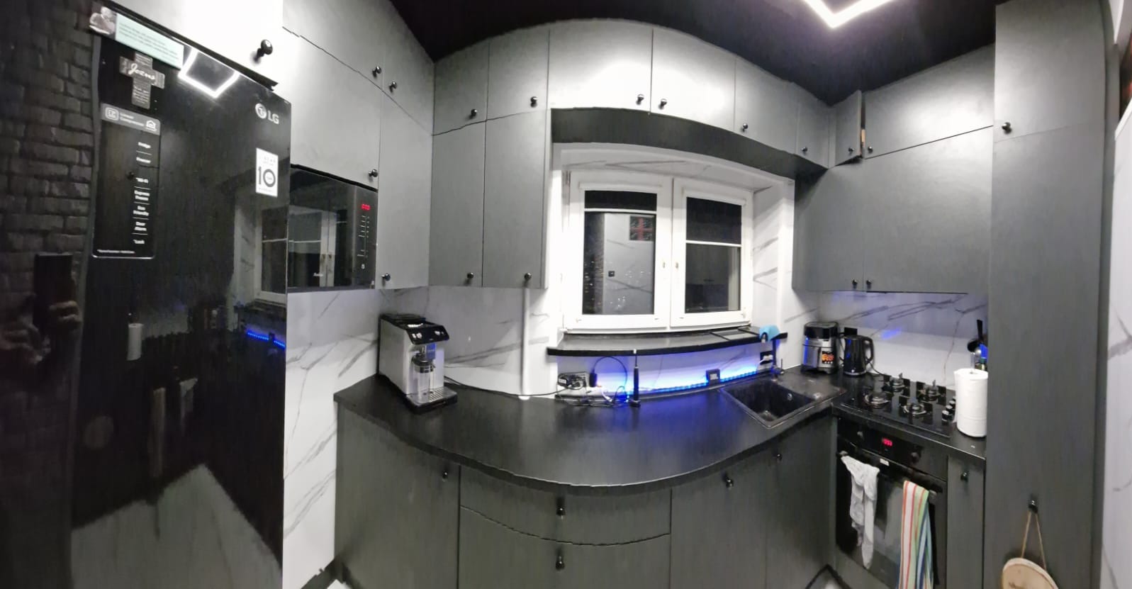 Nowoczesna, szara kuchnia z czarnym sufitem i białym marmurem na ścianach. Widoczny sprzęt AGD: lodówka LG, mikrofalówka, ekspres do kawy. Oświetlenie LED pod szafkami.