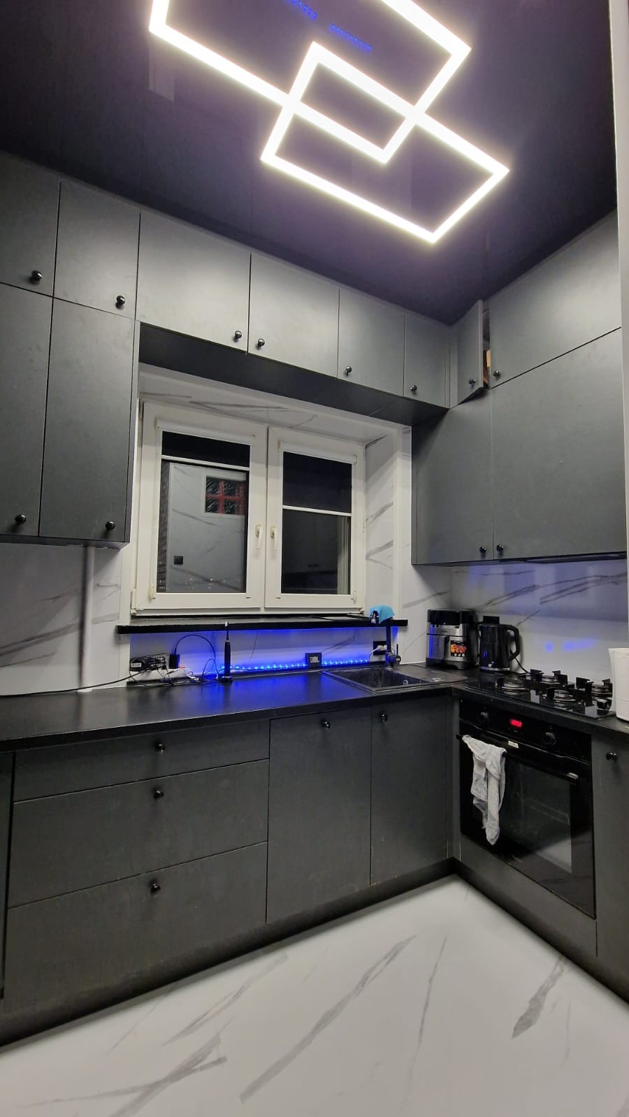Nowoczesna, szara kuchnia z białym blatem i marmurową podłogą. Podświetlenie LED pod szafkami i geometryczna lampa sufitowa dodają unikalnego charakteru.