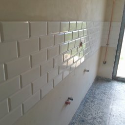 JankoBud - Ściana z białych, błyszczących płytek ułożonych w cegiełkę. Widoczne przyłącza wodne i fragment instalacji miedzianej. Na podłodze wzorzyste, szare płytki.
