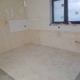 JankoBud - Wnętrze w trakcie remontu: częściowo ułożone białe płytki na ścianie, okno zabezpieczone folią, widoczne instalacje elektryczne i fragment podłogi z płytek.