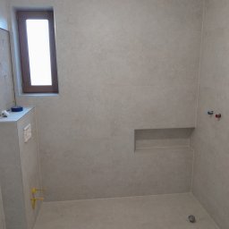 JankoBud - Wykończona łazienka z beżowymi płytkami, wnęką w ścianie i oknem. Widoczne przyłącza wodne i kanalizacyjne. Minimalistyczny design, nowoczesne wykończenie.
