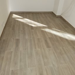 JankoBud - Jasne, drewnopodobne płytki ułożone w pustym pokoju, z widocznym fragmentem ściany i listwą przypodłogową. Na podłodze widoczny pas światła słonecznego.