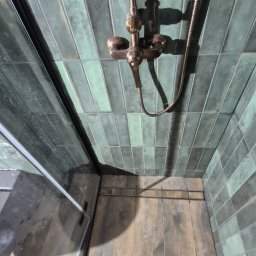 JankoBud - Kabina prysznicowa z zielonymi, podłużnymi płytkami i miedzianą armaturą. Widoczny odpływ liniowy i fragment buta. Ujęcie z góry.