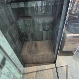 JankoBud - Wykończona kabina prysznicowa z ciemnozielonymi, błyszczącymi płytkami ułożonymi wertykalnie. Widoczna półka wewnątrz i fragment umywalki z szafką. Ujęcie z dołu.