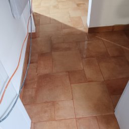 JankoBud - Ułożona, brązowa terakota podłogowa w korytarzu, widok z góry. Widoczne kable przy ścianie i fragment futryny drzwi. Cień podkreśla wzór płytek.