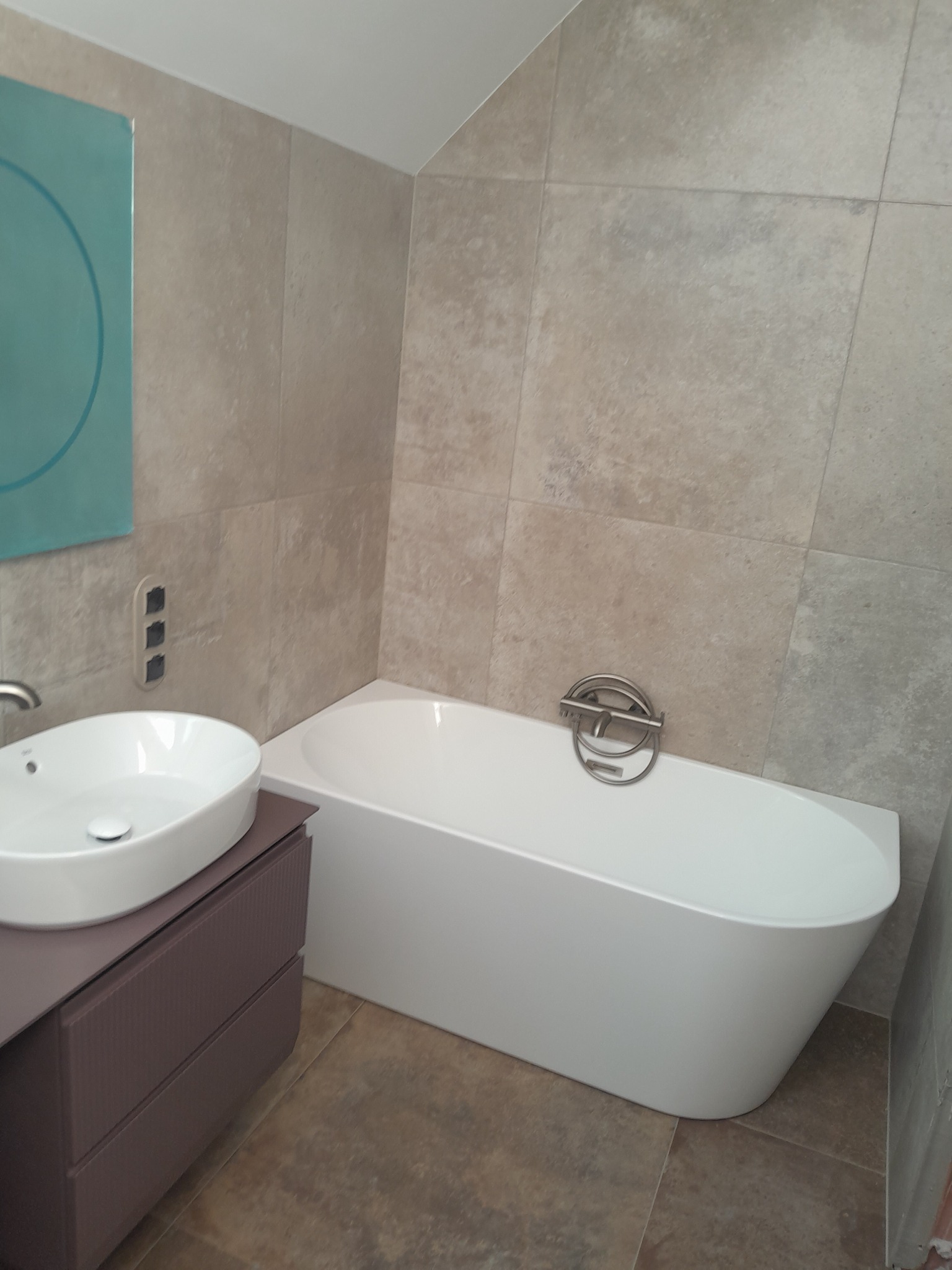 Nowoczesna łazienka z owalną umywalką nablatową na szafce w odcieniach fioletu i białą wanną, wykończona beżowymi płytkami imitującymi kamień.