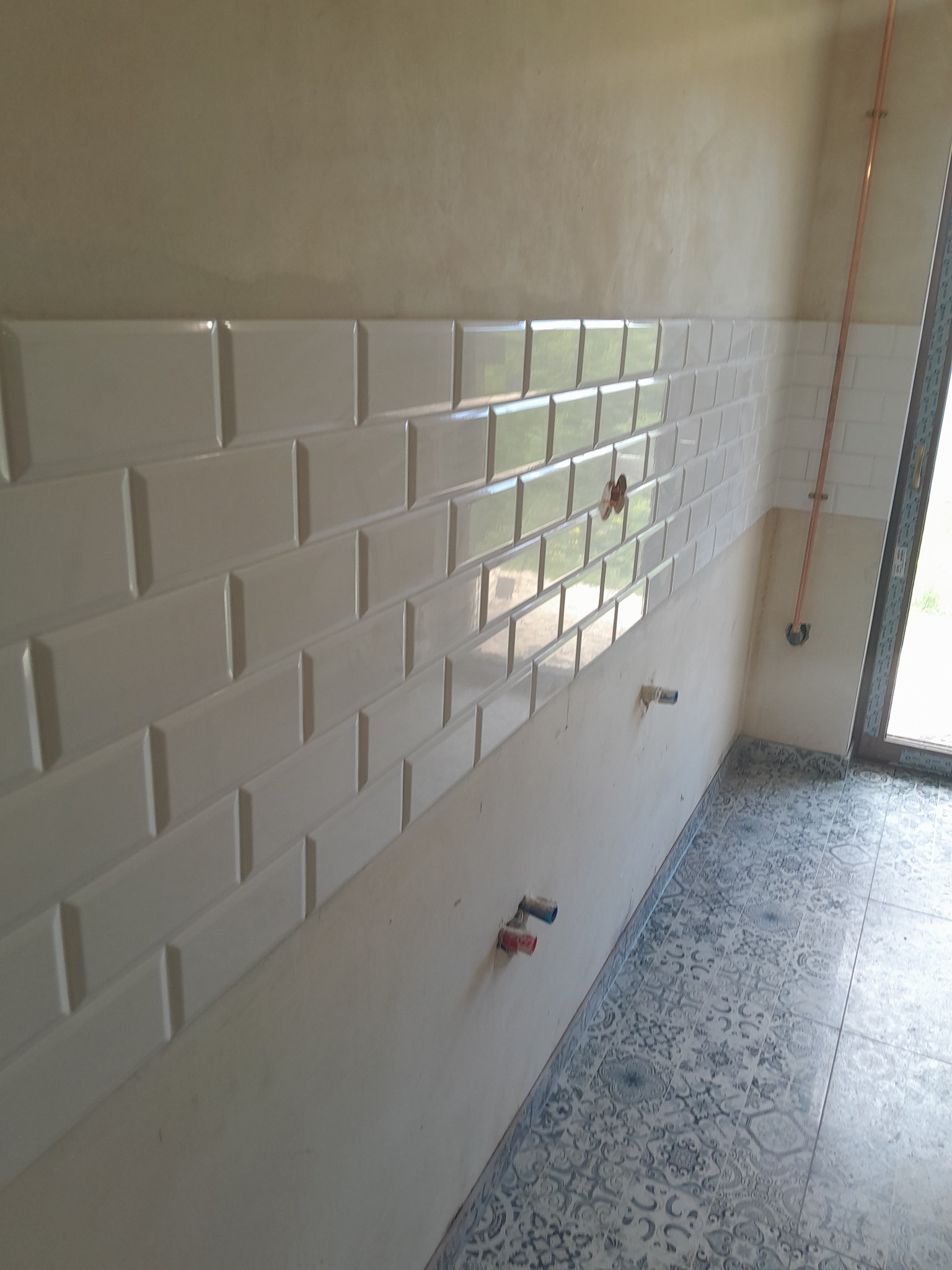 Ściana z białych, błyszczących płytek ułożonych w cegiełkę. Widoczne przyłącza wodne i fragment instalacji miedzianej. Na podłodze wzorzyste, szare płytki.