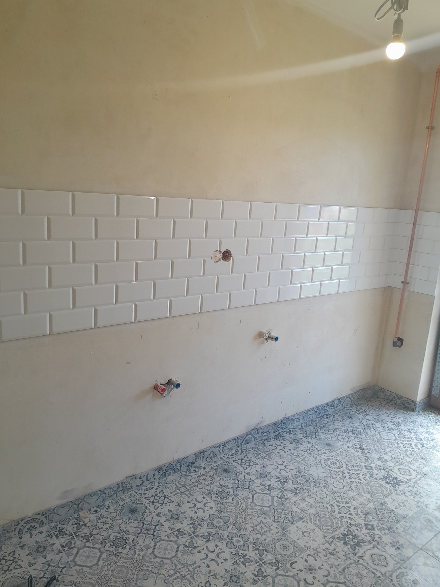 Ściana z ułożonymi białymi, błyszczącymi płytkami typu metro, widoczne instalacje i wzorzysta podłoga. Etap prac wykończeniowych kuchni lub łazienki.