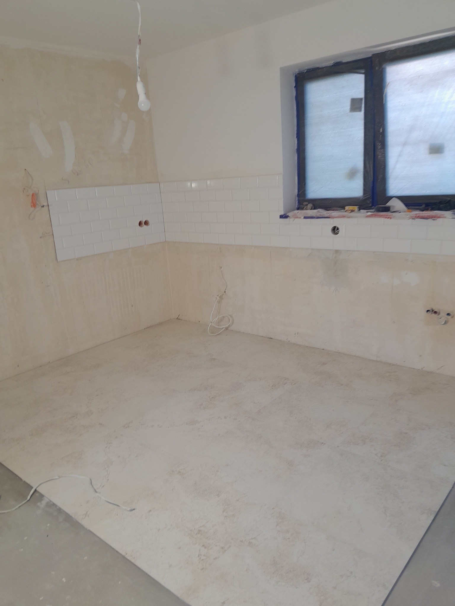 Wnętrze w trakcie remontu: częściowo ułożone białe płytki na ścianie, okno zabezpieczone folią, widoczne instalacje elektryczne i fragment podłogi z płytek.