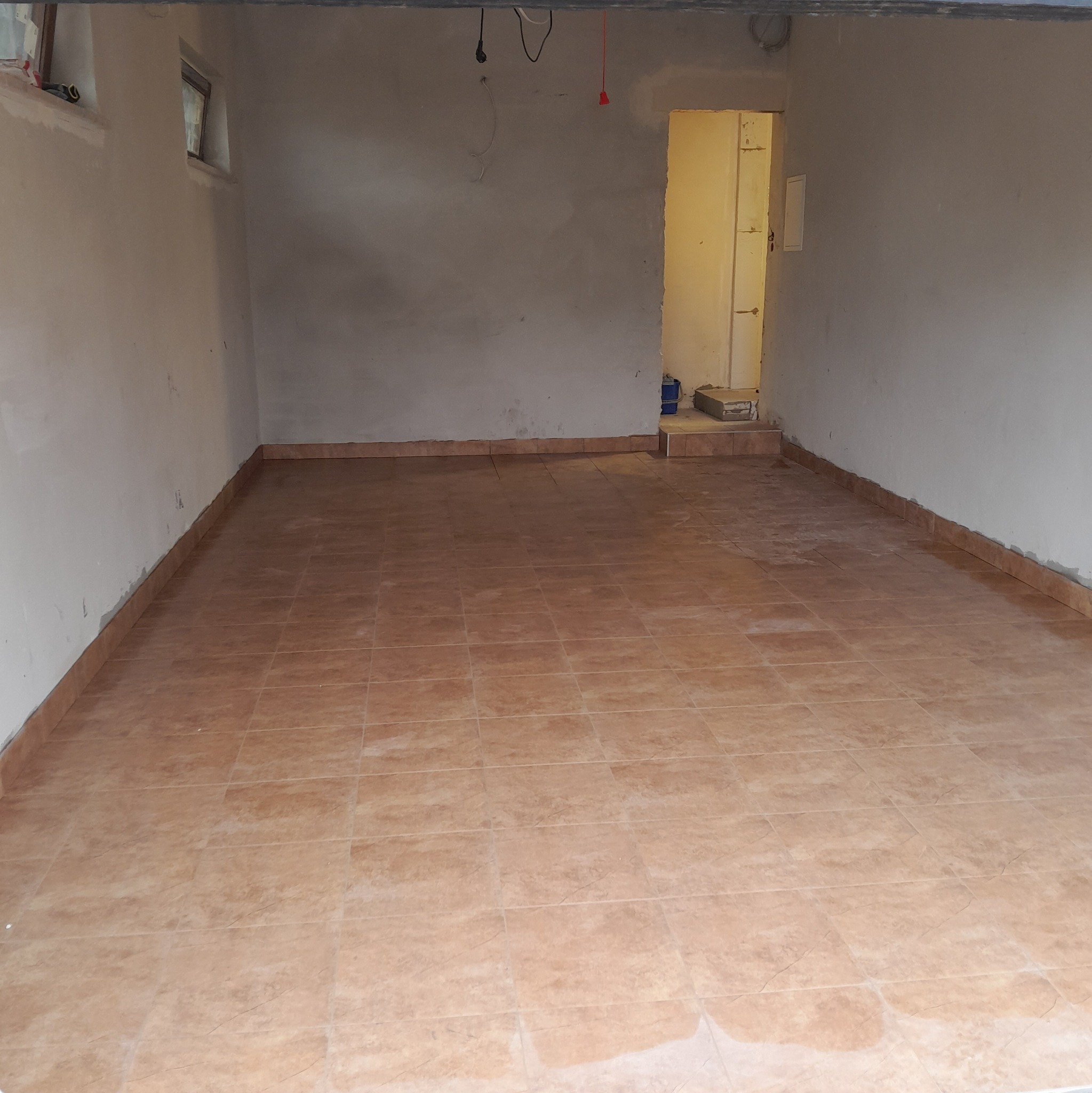 Wyłożona brązowymi płytkami podłoga w surowym pomieszczeniu z oknem, widoczne kable i nieotynkowane ściany. Widok na próg drugiego pomieszczenia.