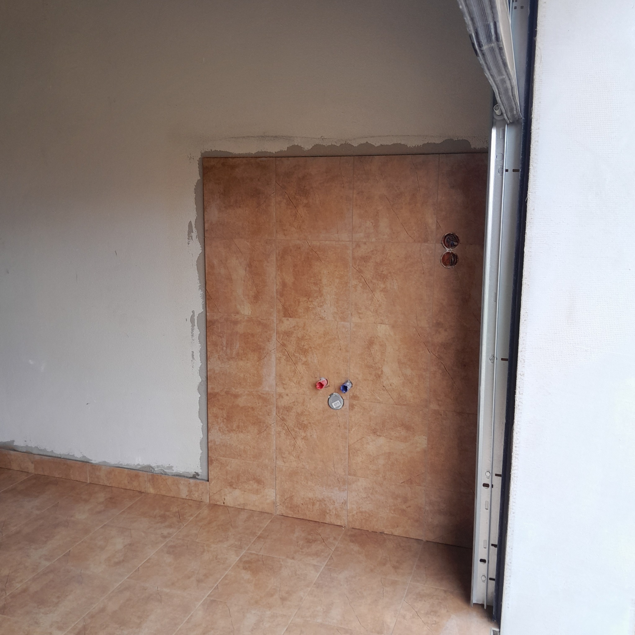 Układanie brązowych płytek imitujących marmur na ścianie i podłodze w trakcie remontu łazienki. Widoczne przyłącza wodne i fragment ramy drzwi.