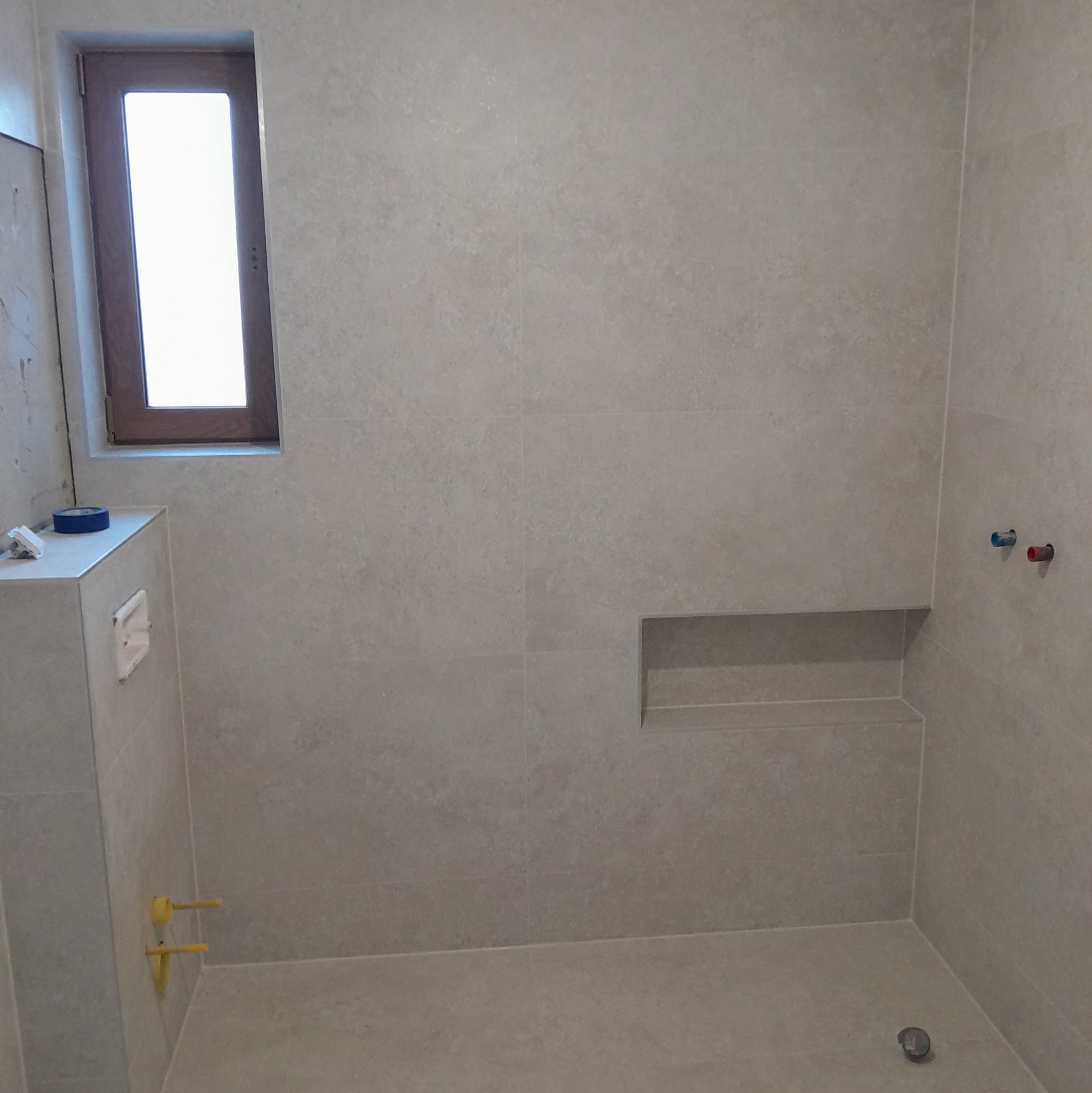 Wykończona łazienka z beżowymi płytkami, wnęką w ścianie i oknem. Widoczne przyłącza wodne i kanalizacyjne. Minimalistyczny design, nowoczesne wykończenie.