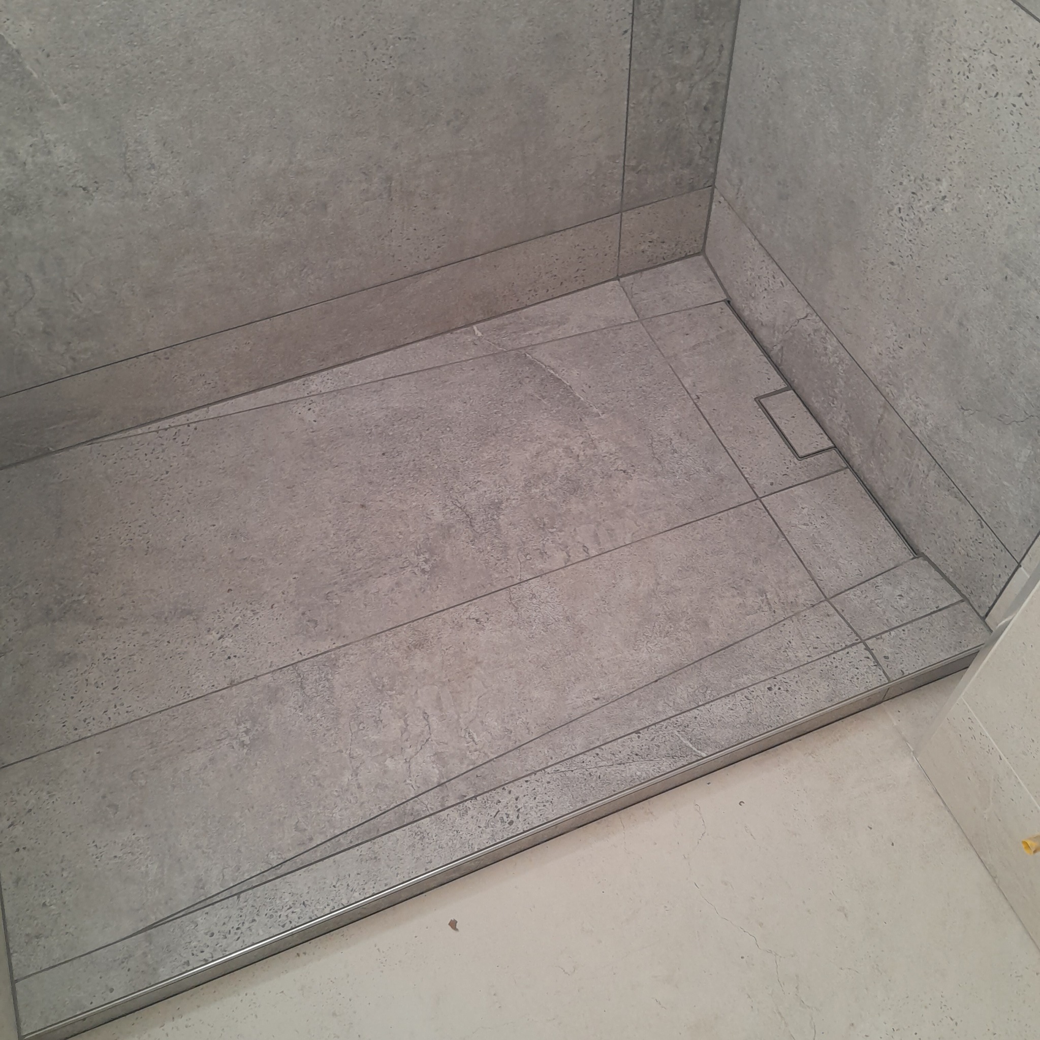 Wykończona strefa prysznicowa z szarymi, wielkoformatowymi płytkami imitującymi kamień, oddzielona metalowym profilem od jasnej posadzki. Widoczny odpływ liniowy.