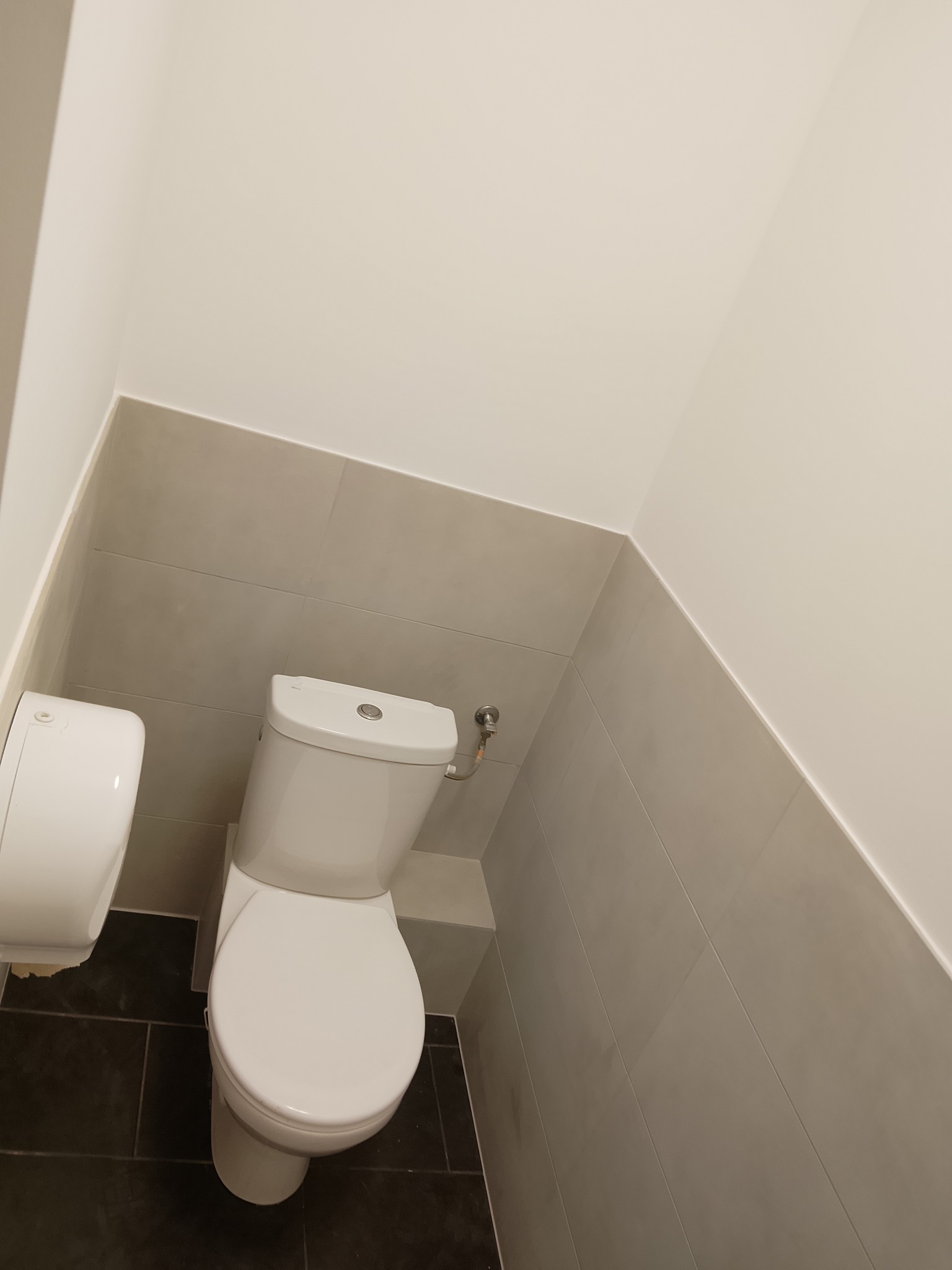 Biała toaleta kompaktowa w rogu wykafelkowanej łazienki z ciemną podłogą i jasnymi płytkami ściennymi. Widoczny podajnik na papier toaletowy.