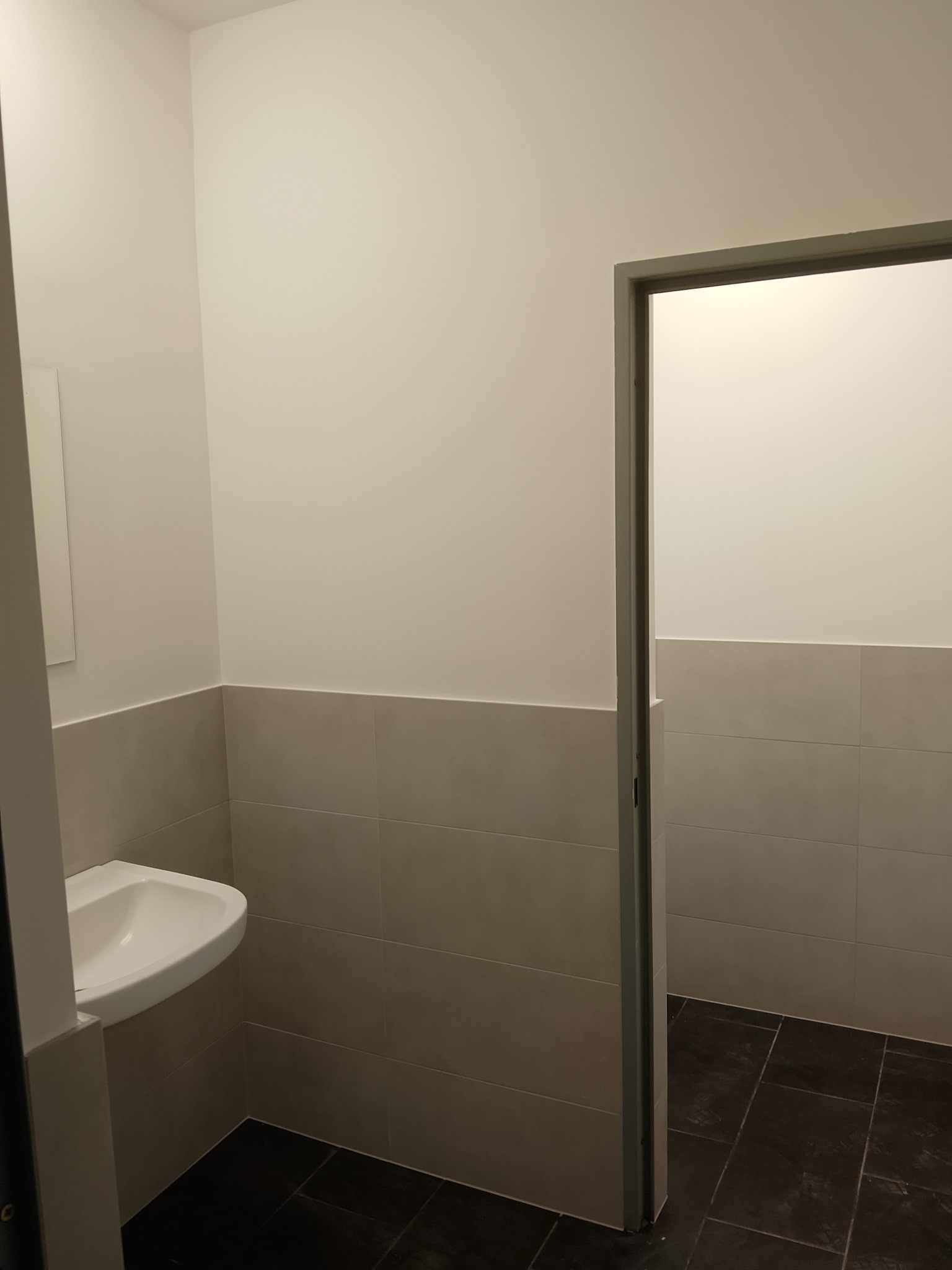 Wykończona łazienka z umywalką i płytkami w odcieniach szarości, widok na otwarte drzwi do drugiego pomieszczenia, czarna podłoga.