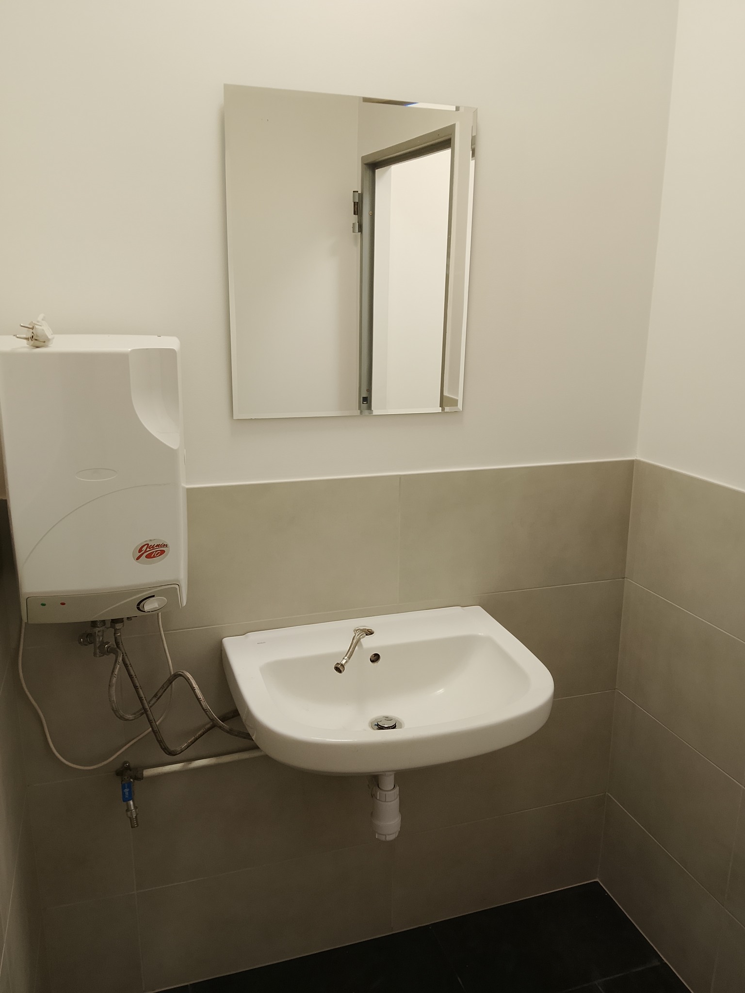 Biała umywalka z baterią i podgrzewaczem wody na ścianie w łazience z szarymi płytkami. W tle lustro i otwarte drzwi.