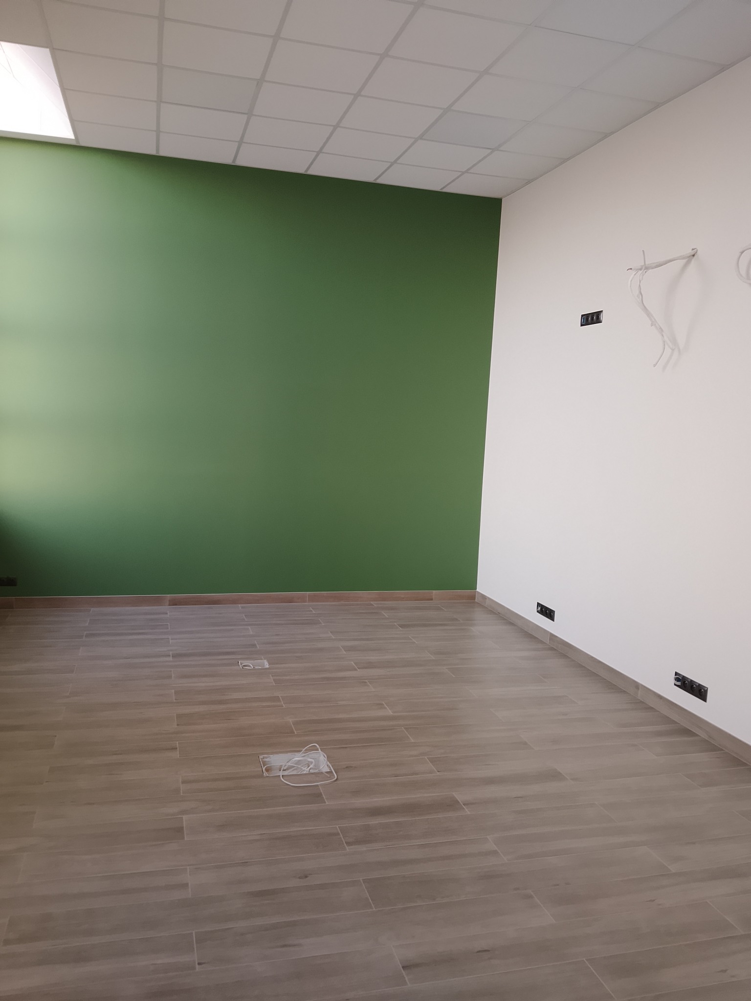 Wnętrze po remoncie: zielona ściana, ułożona podłoga z płytek imitujących drewno, biała ściana z wyprowadzonymi kablami i gniazdkami. Widok na wykończone pomieszczenie.