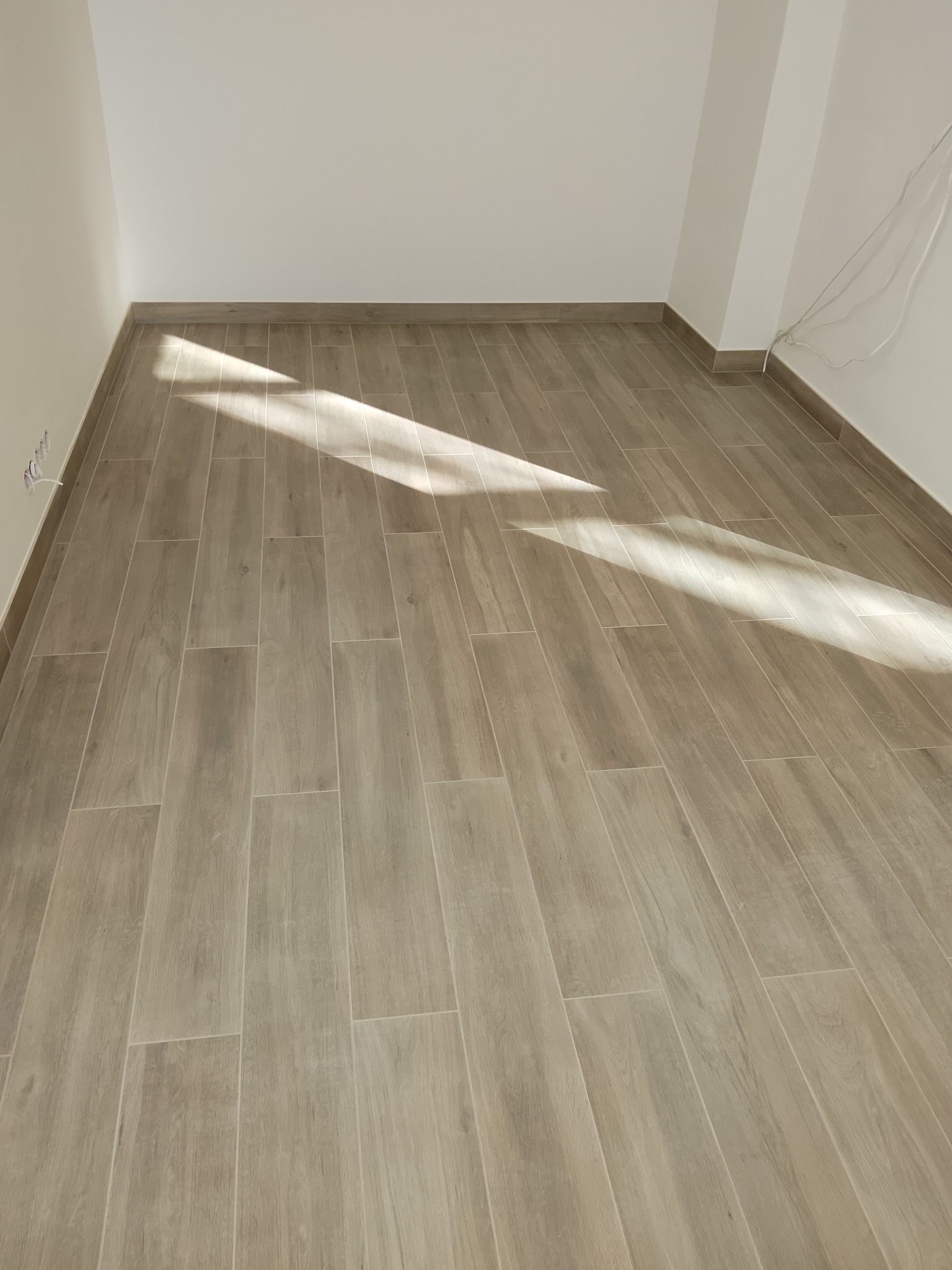 Jasne, drewnopodobne płytki ułożone w pustym pokoju, z widocznym fragmentem ściany i listwą przypodłogową. Na podłodze widoczny pas światła słonecznego.