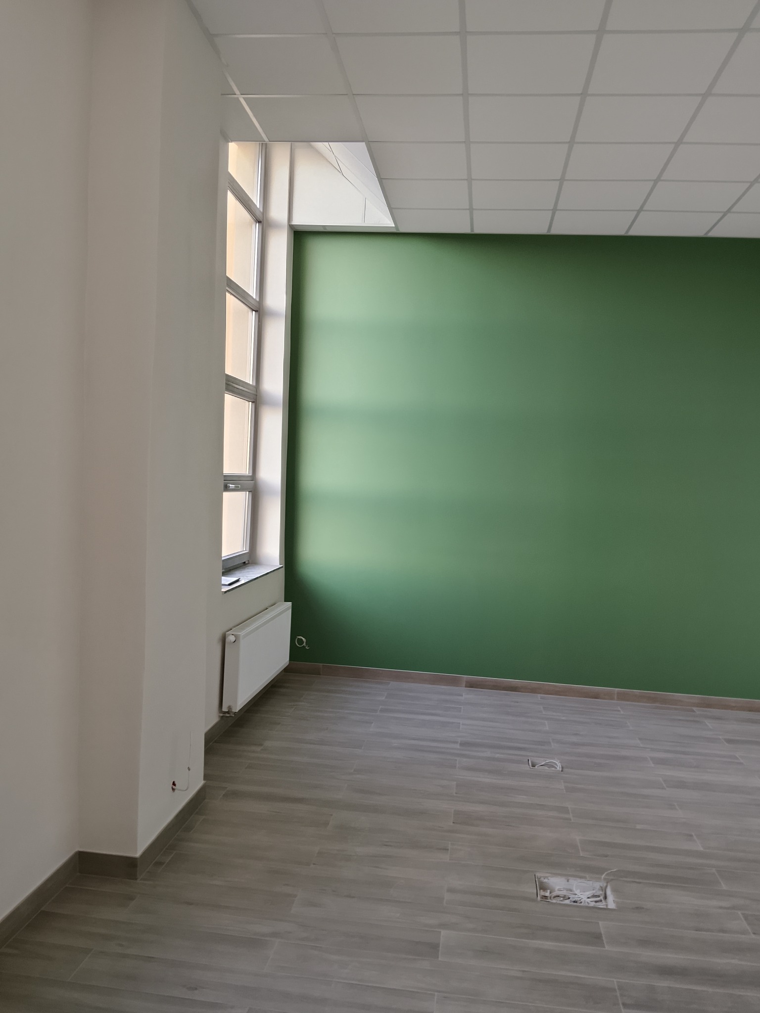 Wykończone wnętrze biura: zielona ściana, szara podłoga z płytek imitujących drewno, białe ściany i sufit podwieszany. Widoczne gniazdka elektryczne w podłodze.